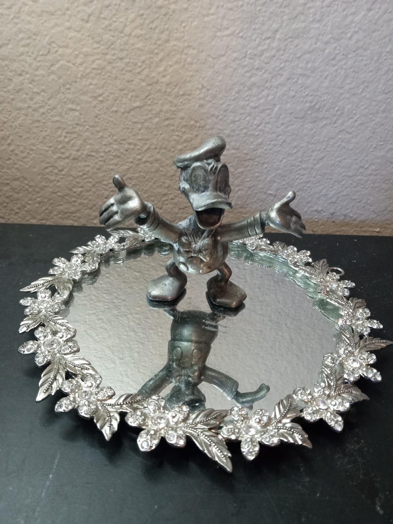 Puede incluir: Una figura plateada de Donald Duck con los brazos extendidos se encuentra sobre una bandeja ovalada con espejo. La bandeja est&aacute; bordeada con un dise&ntilde;o floral adornado con cristales transparentes. El reflejo de la figura es visible en el espejo.
