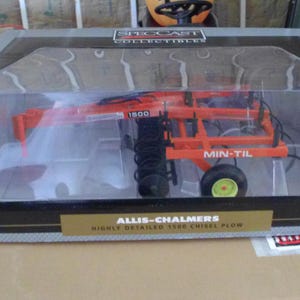 May include: A Speccast Collectibles Allis-Chalmers 1500 Min-Til chisel plow scale model inside a clear plastic display box. The orange metal farm implement features black disc blades, a yellow wheel hub, and white text labels on the frame.