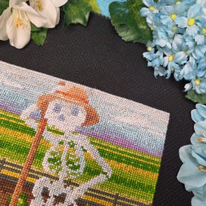 May include: A cross-stitch design featuring a skeleton wearing a straw hat and holding a wooden staff, set against a backdrop of a field and sky. The skeleton is white, the hat is brown, and the field is green and yellow.