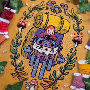 May include: A cross-stitch design featuring a whimsical character riding a backpack, surrounded by mushrooms and foliage. The color palette includes shades of yellow, purple, red, and green. The design is framed by a leafy border and is set against a mustard-colored background.