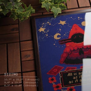 May include: A cross-stitch embroidery framed in a dark wood frame. The design features a red hooded figure with a black bird head against a blue background with yellow stars and a crescent moon. The text "All is as it ever was, as it always will be" is stitched in white on a brown banner.