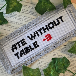 May include: Cross-stitched bookmark with the words "ATE WITHOUT TABLE" in black, with a red graphic. The bookmark has a white background, gray border, and lace trim. The bookmark is on a vintage cookbook page.