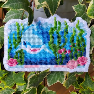 May include: A cross-stitch of an underwater scene. It shows a white shark with a pink mouth, set against blue water, green seaweed, and pink coral. The edges are finished with white thread, creating a framed effect.