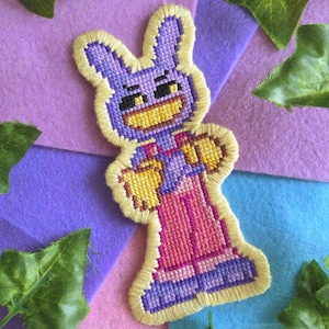 May include: A handmade, embroidered patch featuring a cartoon rabbit character. The rabbit is purple with yellow accents, wearing pink overalls. The patch is outlined in yellow and set against a colorful felt background.
