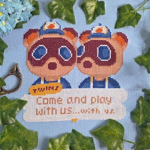 Puede incluir: Obra de arte de punto de cruz con dos personajes parecidos a mapaches con gorras y uniformes azules. El texto dice "TWINS" y "Come and play with us...with us." El fondo es azul claro, rodeado de hojas verdes y flores azules.