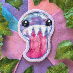 May include: A cross-stitch patch featuring a stylised creature with a round, light purple head, large black eyes, and a wide mouth filled with sharp white teeth. A long, bright pink tongue extends downwards from the mouth, all outlined in light purple thread.