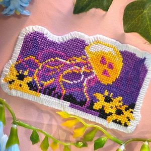 Peut inclure: Un patch de point de croix fait à la main avec un design coloré. Le patch a un fond violet avec des accents jaunes, roses et noirs. Le motif comprend un crâne jaune et une bordure frangée blanche. Le patch est sur une surface rose.