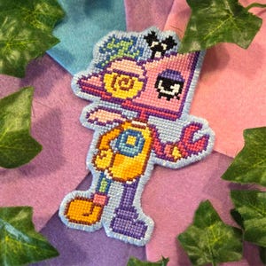Può includere: Un personaggio robotico colorato a punto croce con testa rosa, busto giallo e gambe viola. Il personaggio ha un braccio a forma di chiave inglese ed è delineato in azzurro. Lo sfondo è un mix di tessuto rosa, blu e viola, con foglie verdi.