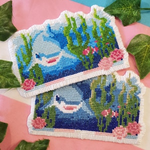 Peut inclure: Deux pièces de point de croix faites à la main représentant une baleine bleue et blanche dans une scène sous-marine. Le motif comprend des algues vertes, des fleurs roses et une bordure blanche. Le fond est un tissu à rayures roses et blanches.