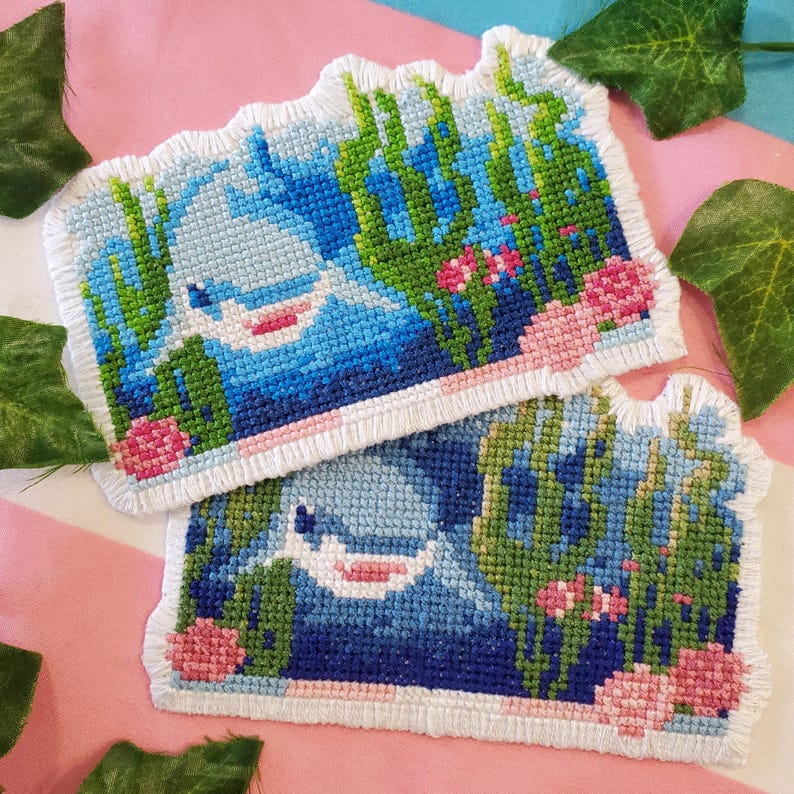 May include: Two handmade cross-stitch pieces depicting a blue and white whale in an underwater scene. The design includes green seaweed, pink flowers, and a white border. The background is a pink and white striped fabric.