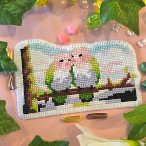 May include: A cross-stitch piece featuring two kissing birds perched on a branch. The birds have pink heads and green wings, set against a sky-blue and pink background. The piece is framed with a white border and rests on a pink surface.