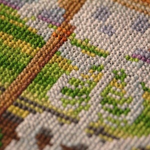 May include: Close-up of a colorful cross-stitch pattern. The design features a mix of green, yellow, brown, white, and purple threads, creating a textured, geometric pattern. The stitches are tightly woven, showcasing the detailed craftsmanship.