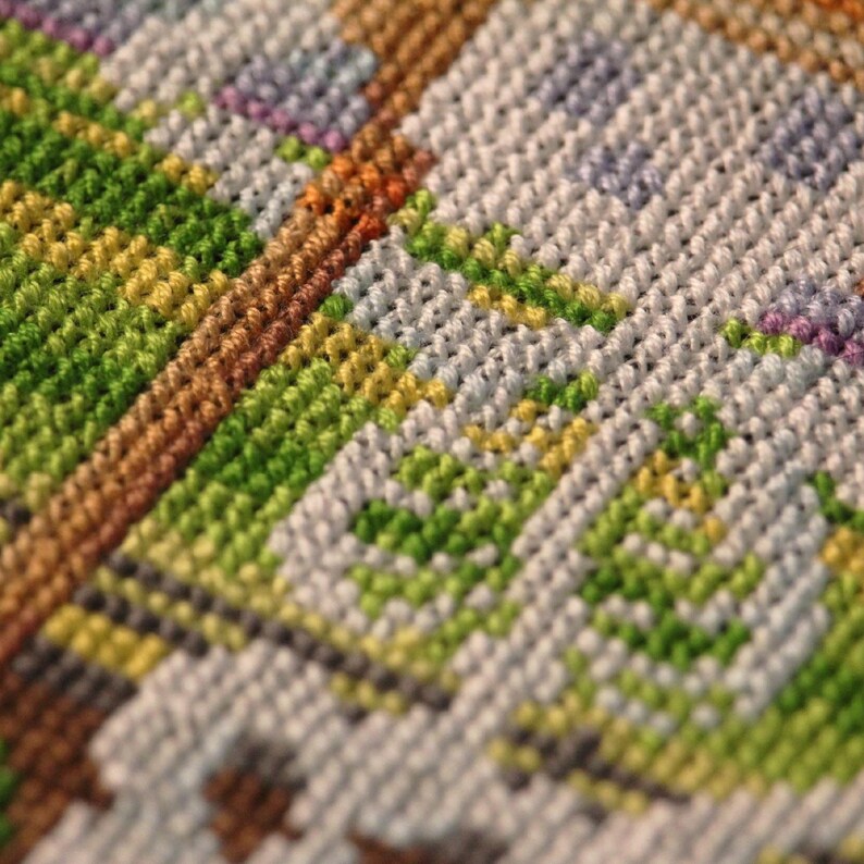 May include: Close-up of a colorful cross-stitch pattern. The design features a mix of green, yellow, brown, white, and purple threads, creating a textured, geometric pattern. The stitches are tightly woven, showcasing the detailed craftsmanship.