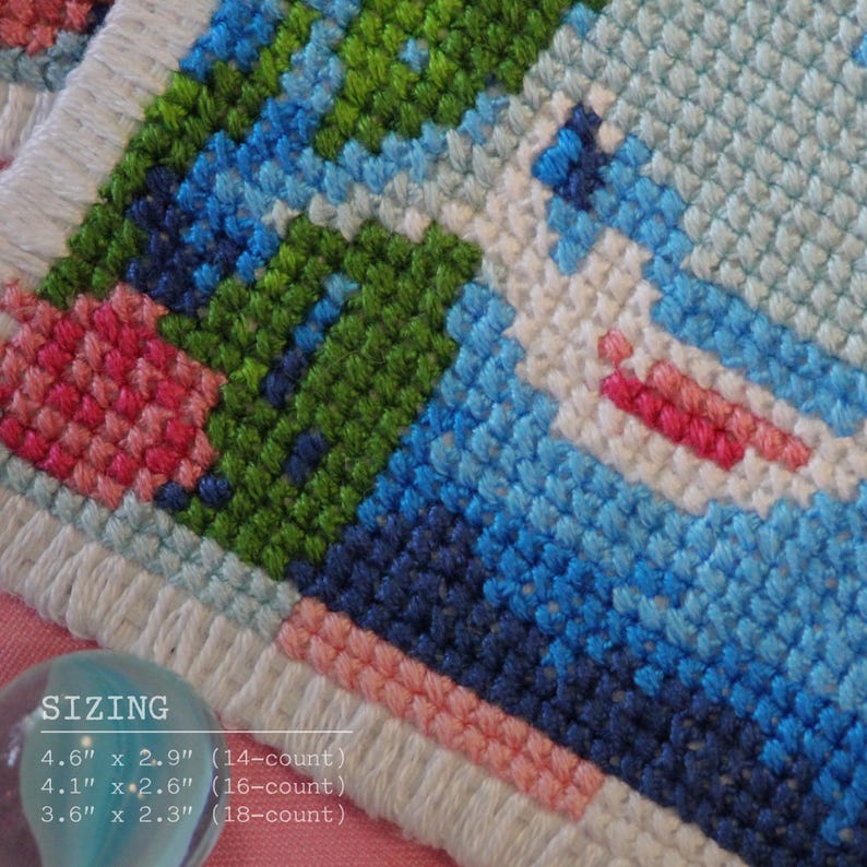 May include: A close-up of a cross-stitch design with a pixelated image in blue, green, pink, and white. The text "SIZING" is visible, along with dimensions: 11.7 cm x 7.4 cm, 10.4 cm x 6.6 cm, and 9.1 cm x 5.8 cm. The cross-stitch is on a white fabric.