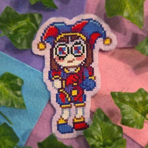 May include: A colourful, pixelated jester patch. The patch features a jester character with a red, blue, and yellow outfit, a jester hat, and glasses. The patch is surrounded by a light purple border. The background includes green leaves and colourful felt.