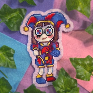 Peut inclure: Un écusson au point de croix représentant un personnage portant un chapeau de bouffon aux accents rouges, bleus et jaunes. La figure a des cheveux bruns, de grands yeux aux détails bleus et rouges, et porte une tenue assortie aux blocs de couleurs.