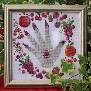 Puede incluir: Una mano blanca con una joya roja en el centro, rodeada de una corona de frutas y bayas rojas, naranjas y moradas. La mano está bordada en punto de cruz y enmarcada en un marco plateado.