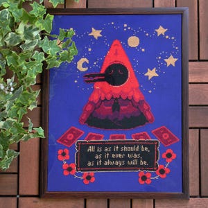 May include: A cross-stitch embroidery framed in a dark wood frame. The design features a hooded figure with a long beak, surrounded by stars and a crescent moon. The text below reads "All is as it should be, as it ever was, as it always will be."