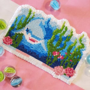May include: A cross-stitch artwork showing an underwater scene. It features a white shark in blue water, with green seaweed and pink coral. The edges are finished with a white fringe, creating a decorative piece.