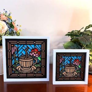 Può includere: Due opere d'arte incorniciate in stile vetrata che raffigurano una tazza di caffè con vapore che ne esce e fiori. Il pezzo più grande è a sinistra e quello più piccolo a destra. Entrambe le opere hanno uno sfondo blu con accenti marroni, rossi, rosa e verdi.