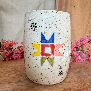 May include: A cylindrical ceramic tumbler with a speckled off-white glaze. The front features a geometric star design composed of a central red square surrounded by blue, yellow, green, and coral triangular points, with small black dots.