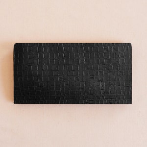 May include: A rectangular, black, textured object with a crocodile skin pattern. The object is centered on a light peach-colored background. The texture appears to be embossed or stamped into the material, creating a tactile surface.
