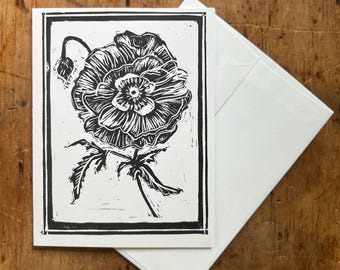 May include: A black and white linocut print of a single poppy flower with a black frame. The print is on a white card.