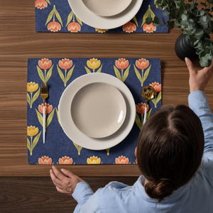 May include: A table setting featuring two navy blue placemats with a tulip pattern in orange, yellow, and green. Each place setting includes a white plate and a gold-colored fork and spoon. The table is made of wood.