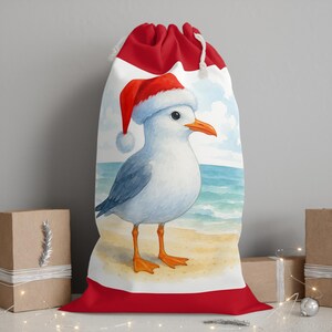 May include: A white drawstring gift bag with a red top and bottom border. The bag features a watercolor illustration of a seagull wearing a Santa hat on a beach. The seagull has blue-grey wings, orange feet and beak, and is standing on a sandy beach.