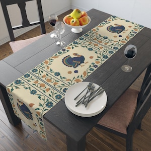 Puede incluir: Camino de mesa color crema con un patrón repetido de un diseño de pavo azul y verde azulado con detalles florales. El camino de mesa está colocado sobre una mesa de madera oscura, con platos, cubiertos y copas de vino.