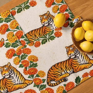 May include: A cream-coloured table runner with a repeating pattern of orange marigold flowers and detailed tiger illustrations. The runner has a border of green leaves and flowers. Lemons are in a basket.