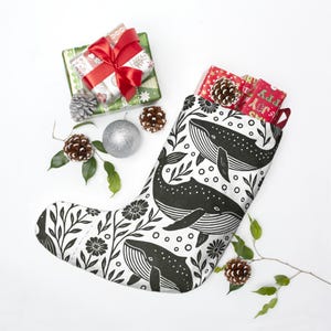 Puede incluir: Un calcetín navideño con un diseño de ballenas en blanco y negro, lleno de regalos envueltos y piñas. El calcetín presenta un diseño repetido de ballenas, flores y hojas. Regalos y adornos adicionales están dispuestos alrededor del calcetín.