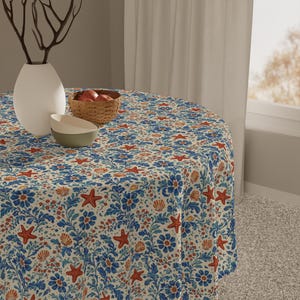 May include: A round tablecloth with a marine-themed pattern. The design features blue floral elements, red starfish, and orange seashells on a cream background. A white vase, a basket of apples, and a bowl are on the table.