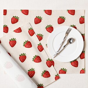 Könnte beinhalten: Ein weißer Teller mit Gabel und Löffel sowie zwei Tischsets mit Erdbeermuster. Die Tischsets haben ein sich wiederholendes Muster aus roten Erdbeeren mit grünen Blättern auf beigem Hintergrund. Ein Tischset ist aufgerollt.