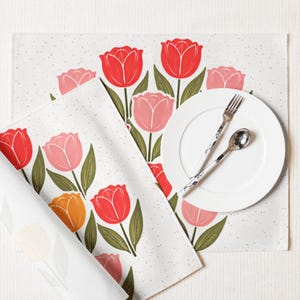 May include: A white placemat and napkin set featuring a tulip design. The tulips are red, pink, and orange with green stems and leaves. A white plate, fork, and spoon are also present.