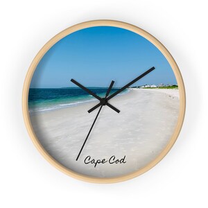 May include: A round wooden wall clock with a beach scene. The clock face features a white sandy beach with blue ocean water and a clear blue sky. The clock hands are black and the clock face has the text "Cape Cod" written in black cursive.