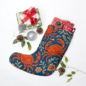 May include: A blue Christmas stocking with a crab pattern, filled with wrapped gifts and pine cones. The stocking has a repeating design of orange crabs and floral elements. Presents are wrapped in green and red paper with red ribbons.