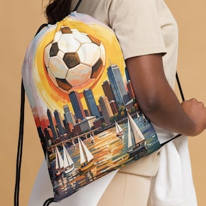 Könnte beinhalten: Kordelzugrucksack mit einem Fußball-Design und einer Stadtlandschaft. Das Kunstwerk zeigt einen großen Fußball, eine Skyline von Gebäuden, Segelboote auf dem Wasser und einen lebendigen Sonnenuntergang. Die Tasche hat schwarze Kordelzüge.