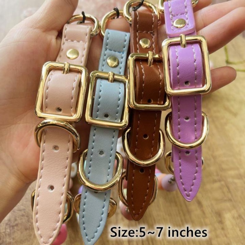 May include: A collection of four colorful dog collars in pink, blue, brown, and purple. Each collar features a gold-colored buckle and hardware. The collars are approximately 5 to 7 inches in length.