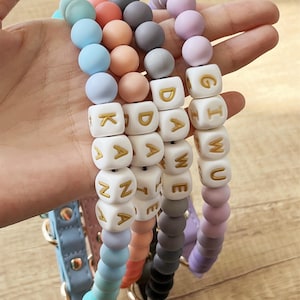 Può includere: Una collezione di collari per cani con perline colorate. I collari presentano una varietà di perline color pastello e perline a cubo bianche con lettere dorate che compongono nomi. I collari hanno cinturini in pelle di diversi colori e finiture dorate.