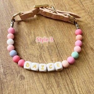 May include: A pink dog collar with the word "DAICE" spelled out in white letter beads. The collar features a pink leather strap with gold-colored hardware and is adorned with colorful round beads in shades of pink, white, and blue.