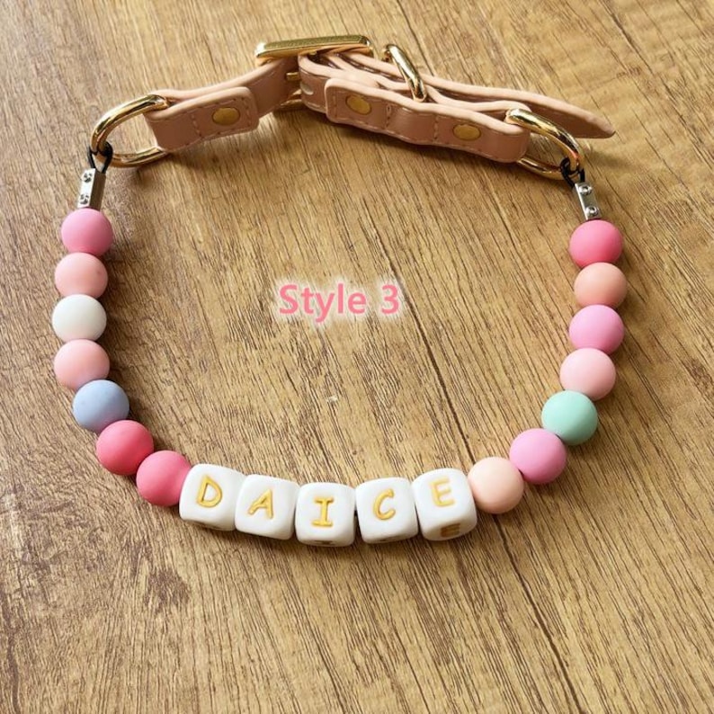 May include: A pink dog collar with the word "DAICE" spelled out in white letter beads. The collar features a pink leather strap with gold-colored hardware and is adorned with colorful round beads in shades of pink, white, and blue.