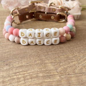 May include: A dog collar with the word "DANCE" spelled out in white letter beads. The collar features a pink leather strap and gold-colored hardware. The beads are in shades of pink, peach, and blue.
