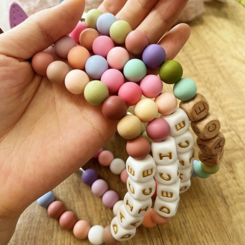 May include: A collection of colorful, round beads in various pastel shades, including pink, blue, green, and brown. Some strands feature white lettered cubes and wooden cubes. The beads are held in a hand, showcasing the variety of colors and textures.