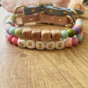 May include: Two dog collars with pastel-colored silicone beads and wooden letter beads spelling "NACE" and "DAICE". The collars have leather straps in pink and blue-grey, with gold-colored hardware.