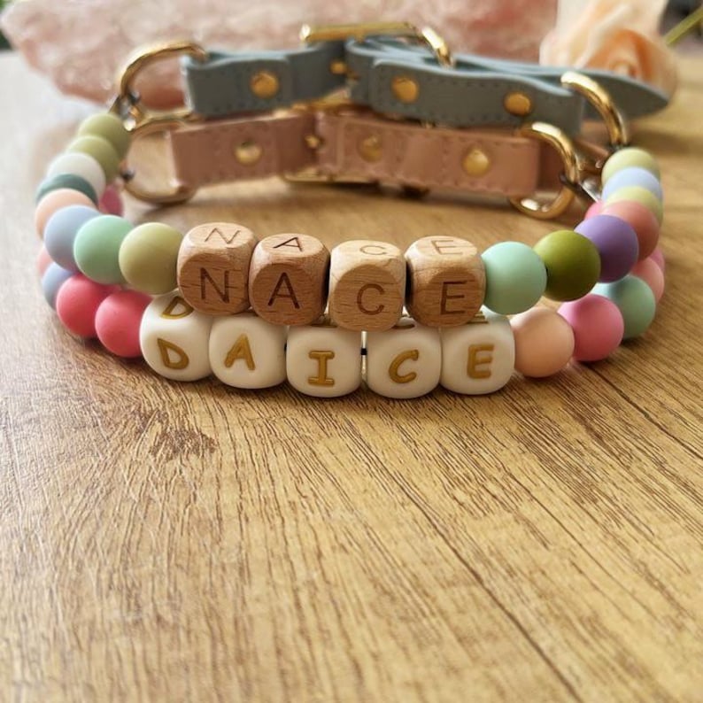 May include: Two dog collars with pastel-colored silicone beads and wooden letter beads spelling "NACE" and "DAICE". The collars have leather straps in pink and blue-grey, with gold-colored hardware.