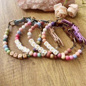 May include: A collection of colorful beaded dog collars with gold-tone hardware. Each collar features a different name spelled out in white lettered wooden cubes. The collars are in shades of pink, purple, green, and brown.