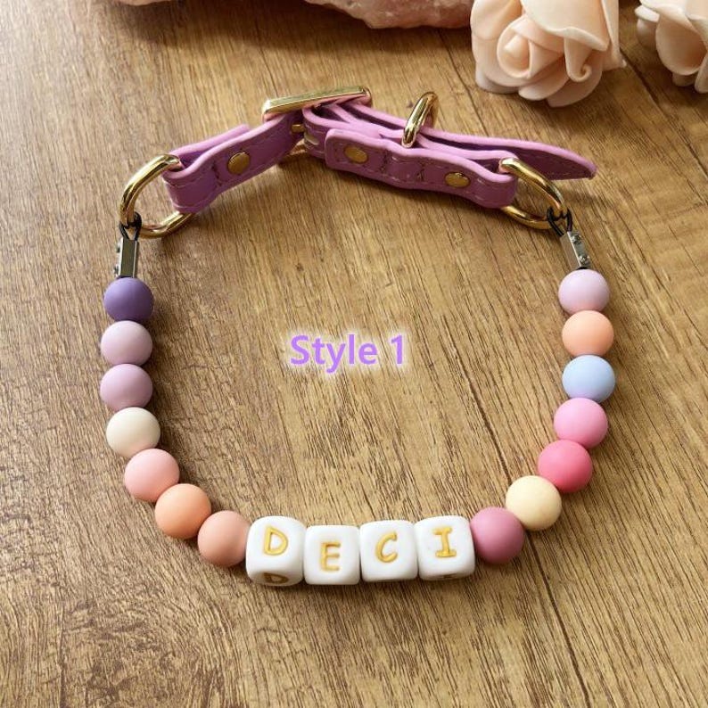 May include: A lavender dog collar with gold-colored hardware and a beaded design. The collar features the name "DECI" spelled out in white letter beads. The beads are in pastel colors, including pink, orange, and purple. The text "Style 1" is also visible.