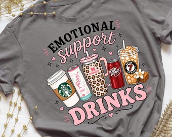 May include: A charcoal grey short-sleeve t-shirt featuring a graphic print with the text "EMOTIONAL SUPPORT DRINKS" in black and pink. The design includes illustrations of a coffee cup, a slim beverage can, a leopard print tumbler, a soda can, and an iced coffee.