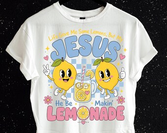 May include: A white t-shirt featuring a graphic print of two yellow cartoon lemons and a glass of lemonade. The text reads: Life Gave Me Some Lemons, But My JESUS He Be Makin' LEMONADE. The design includes blue, pink, and yellow accents.